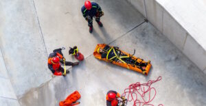 one person secured in a stretcher on the ground, with ropes on the ground, and rescue workers around them wearing safety equipment